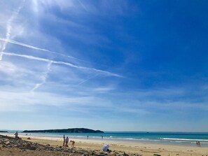 A beach with a clear sky, rocky foreground, and distant hills.