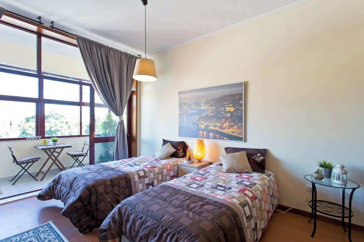 A bedroom with two beds, a window with curtains, a table with chairs, and a painting on the wall.