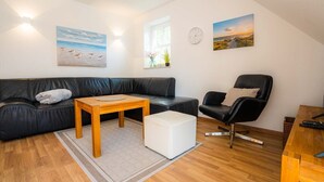 Ein modernes Wohnzimmer mit einem schwarzen Ledersofa, einem Holztisch, einem weißen Hocker, einem schwarzen Ledersessel und einem Beistelltisch aus Holz. An den Wänden hängen zwei Gemälde, und es gibt eine Uhr sowie ein Fenster mit Blick ins Freie.