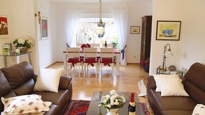 Una sala de estar con dos sofás marrones, una mesa de centro de vidrio con una botella y flores, una zona de comedor con una mesa blanca y sillas rojas, y una araña.