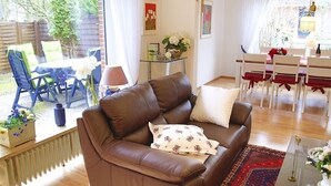Un salón con un sofá de cuero marrón, una mesa de centro de vidrio y una zona de comedor con sillas blancas y mantel rojo.