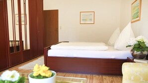 Un dormitorio con cama, armario de madera, almohadas blancas, una mesita con un jarrón de flores y un cuadro en la pared.