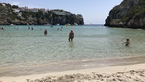 A beach with clear water, people swimming, and boats in the distance. - Modern apartment with a fantastic view over the bay (Cala en Porter)