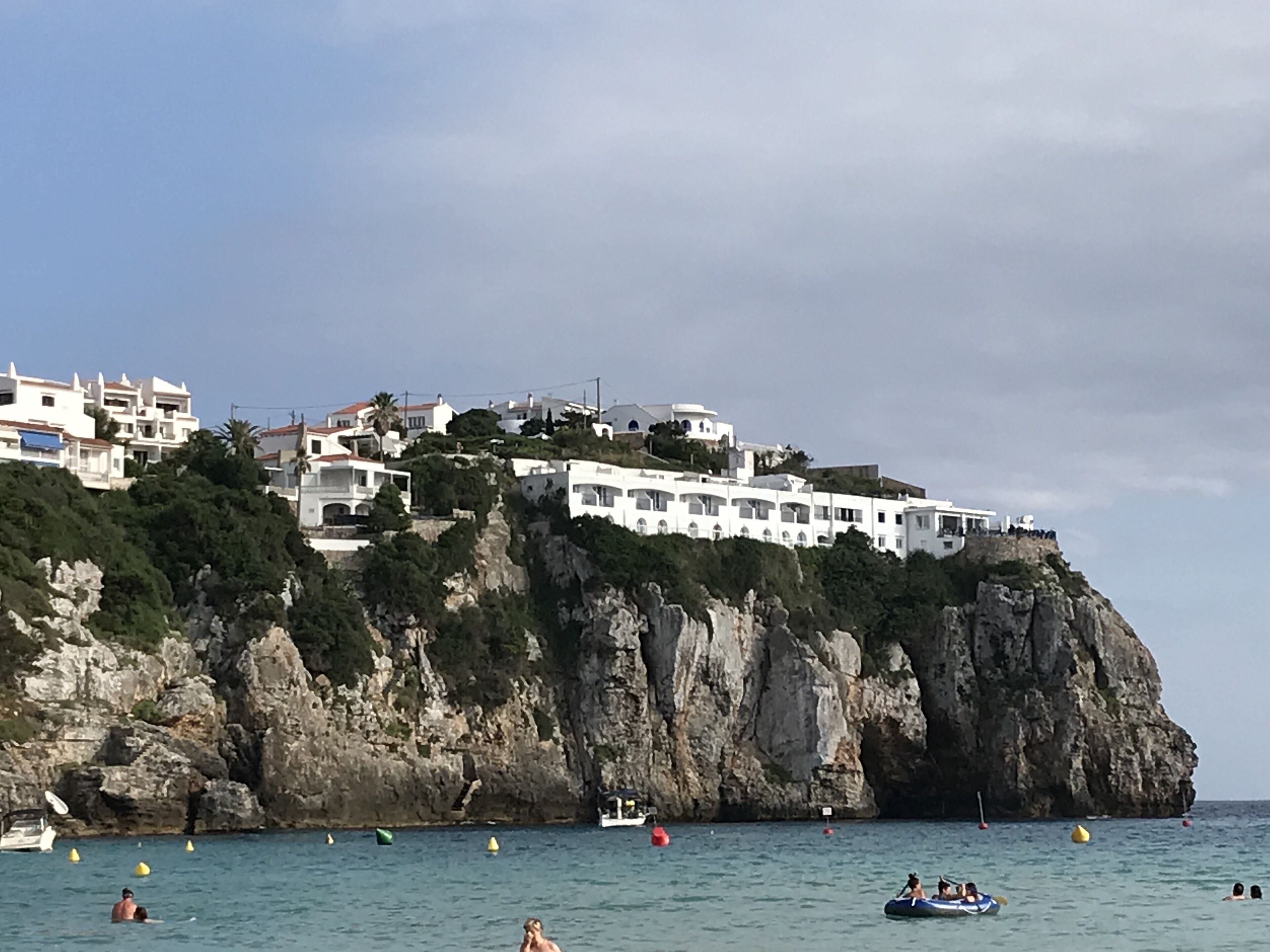 A coastal town with white buildings on a cliff, boats in the water, and people swimming.