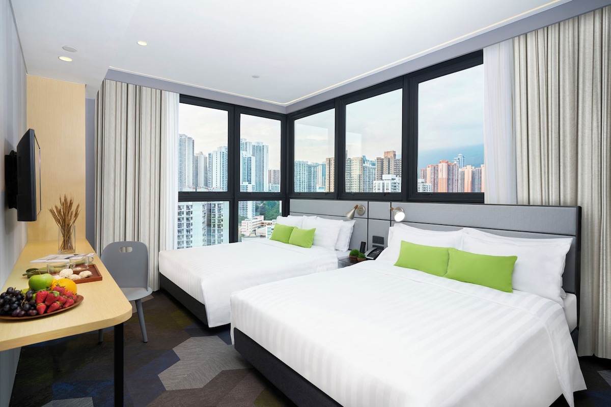 Une chambre d’hôtel avec deux lits, un bureau avec des fruits et une vue sur la ville.