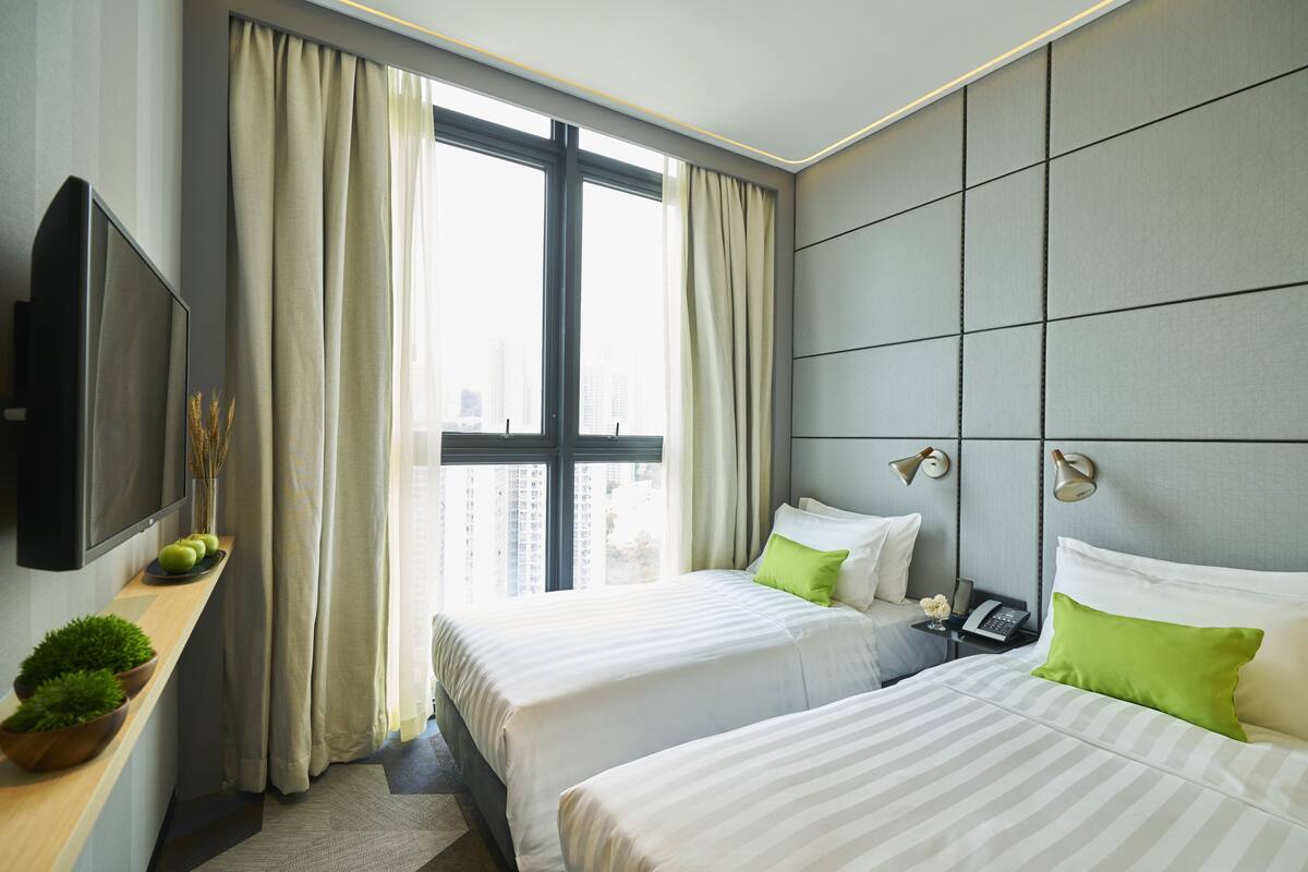 Une chambre d’hôtel avec deux lits, une télévision, un bureau avec une plante et une grande fenêtre avec des rideaux.