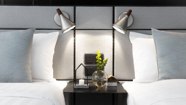 Una cama bien hecha con ropa blanca, una mesita de noche con un florero de plantas verdes y dos lámparas fijadas a la pared.