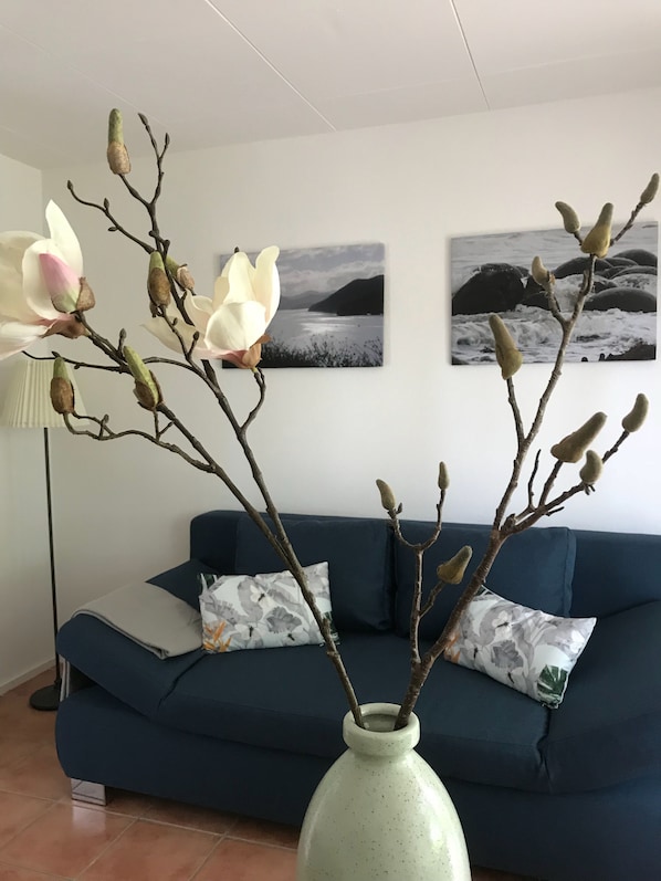 A living room with a blue sofa, a vase with branches and magnolia flowers, and two framed pictures on the wall.