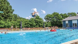 Een zwembad met mensen in en rond het water, een badmeestertoren en een gebouw met een blauw dak op de achtergrond.