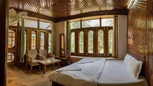 A wooden room with a bed, a chair, a table, and large windows. - Parvati Kuteer (Bhuntar)