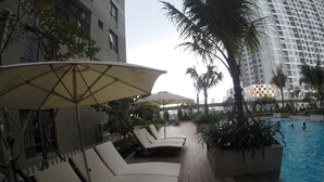 A poolside area with lounge chairs and umbrellas, surrounded by palm trees and a high-rise building in the background. - Riverfront Osis Resort Balcony (Ho Chi Minh City)