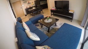 A modern living room with a blue sofa, a coffee table with a vase, a TV, and a dining area with chairs and a table. - Riverfront Osis Resort Balcony (Ho Chi Minh City)