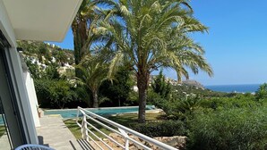 Ein Balkon mit zwei weißen Korbstühlen, einer mit einem grünen Kissen und der andere mit einem gemusterten Kissen. Ein weißes Geländer trennt den Balkon von einem Swimmingpool und üppiger Bepflanzung.