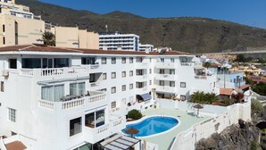A multi-story hotel with balconies, a swimming pool, and a view of a hilly landscape. - Over the Ocean with pool (Candelaria)
