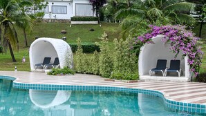 A swimming pool with two covered seating areas, surrounded by greenery and palm trees. - Swiss Valley Hip Resort (Suan Phueng)