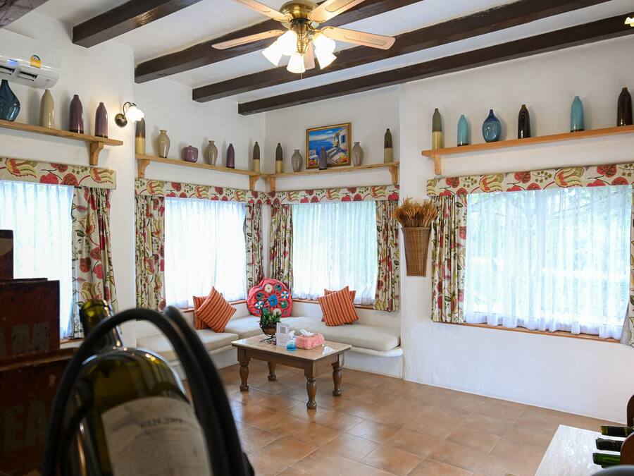Una sala de estar con un sofá, una mesa de centro y un ventilador de techo. Se observan estantes con objetos decorativos y una ventana con cortinas.