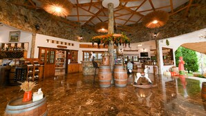 A spacious lobby with a high ceiling, stone walls, wooden beams, and a polished marble floor. There are wooden barrels, a central tree with hanging decorations, and a bar area with bottles and glasses. - Swiss Valley Hip Resort (Suan Phueng)