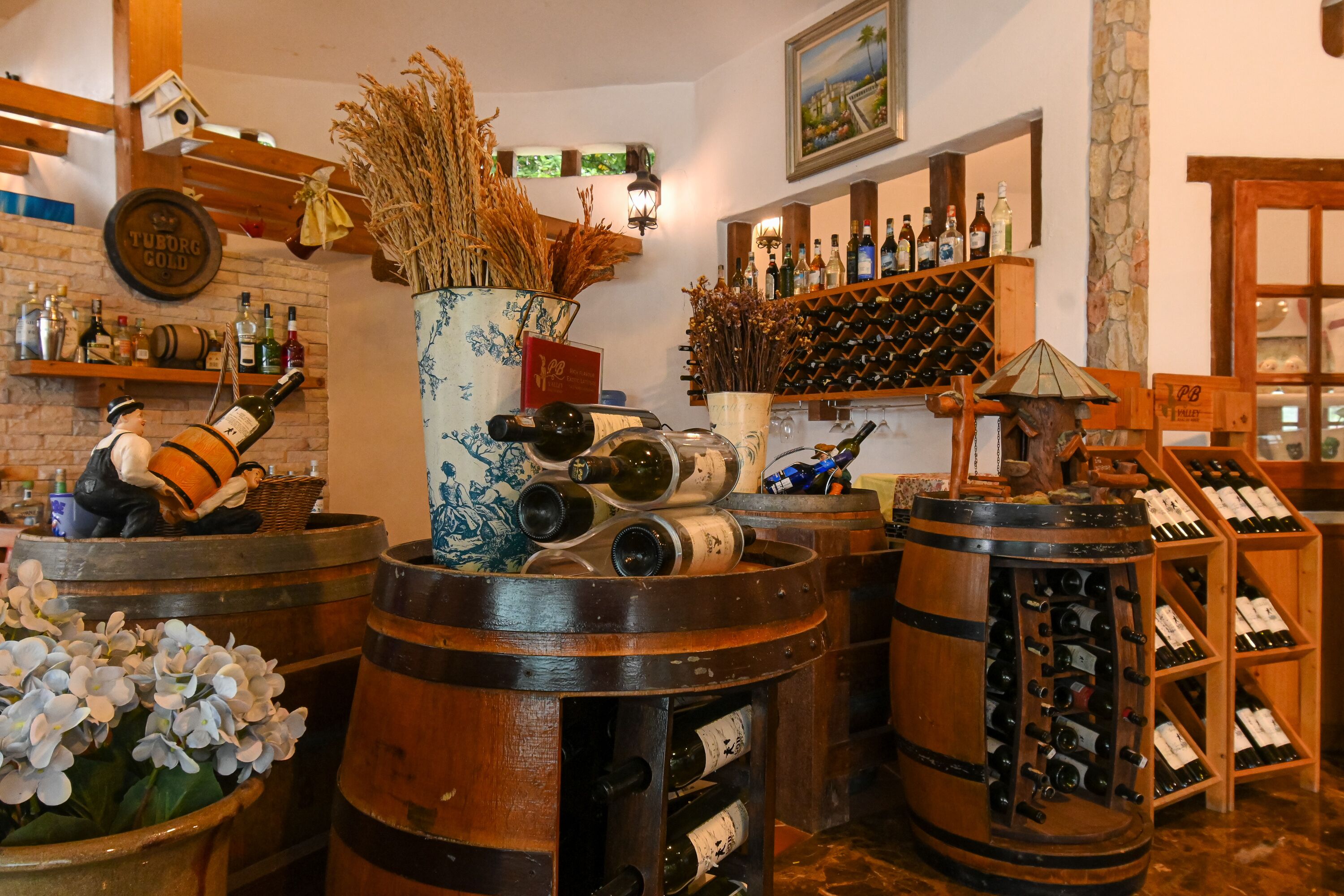Een bar met houten vaten, een grote vaas met gedroogde planten en wijnflessen op planken.