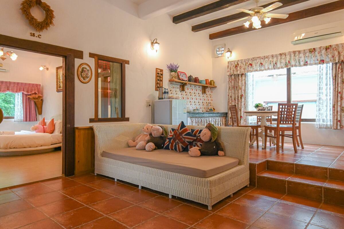 A cozy living room with a wicker sofa, a dining table with chairs, and a kitchen area with a refrigerator and shelves.