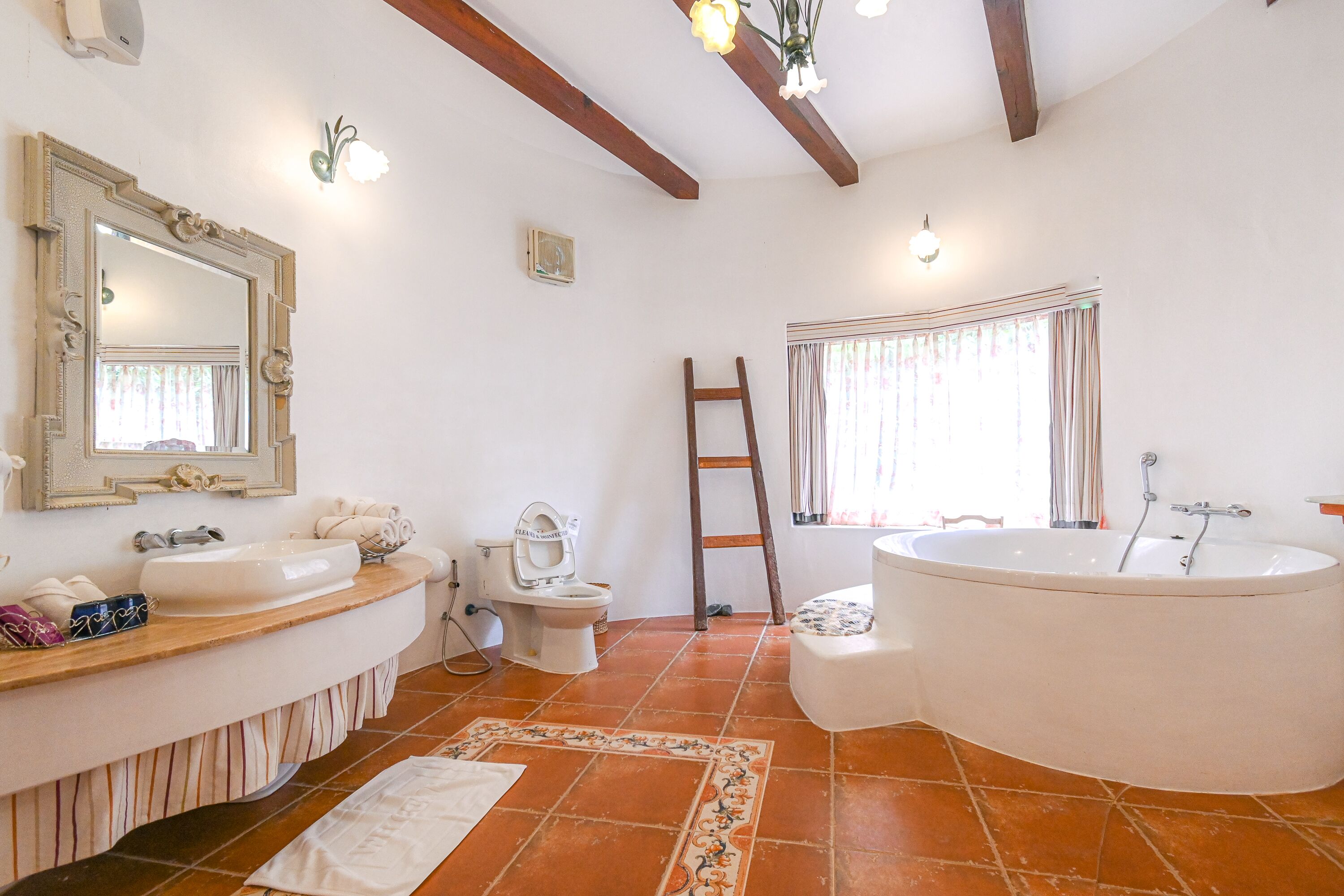 Un baño con una bañera grande, un lavamanos, un inodoro y un espejo. Se ve una escalera apoyada en la pared y una ventana con cortinas.