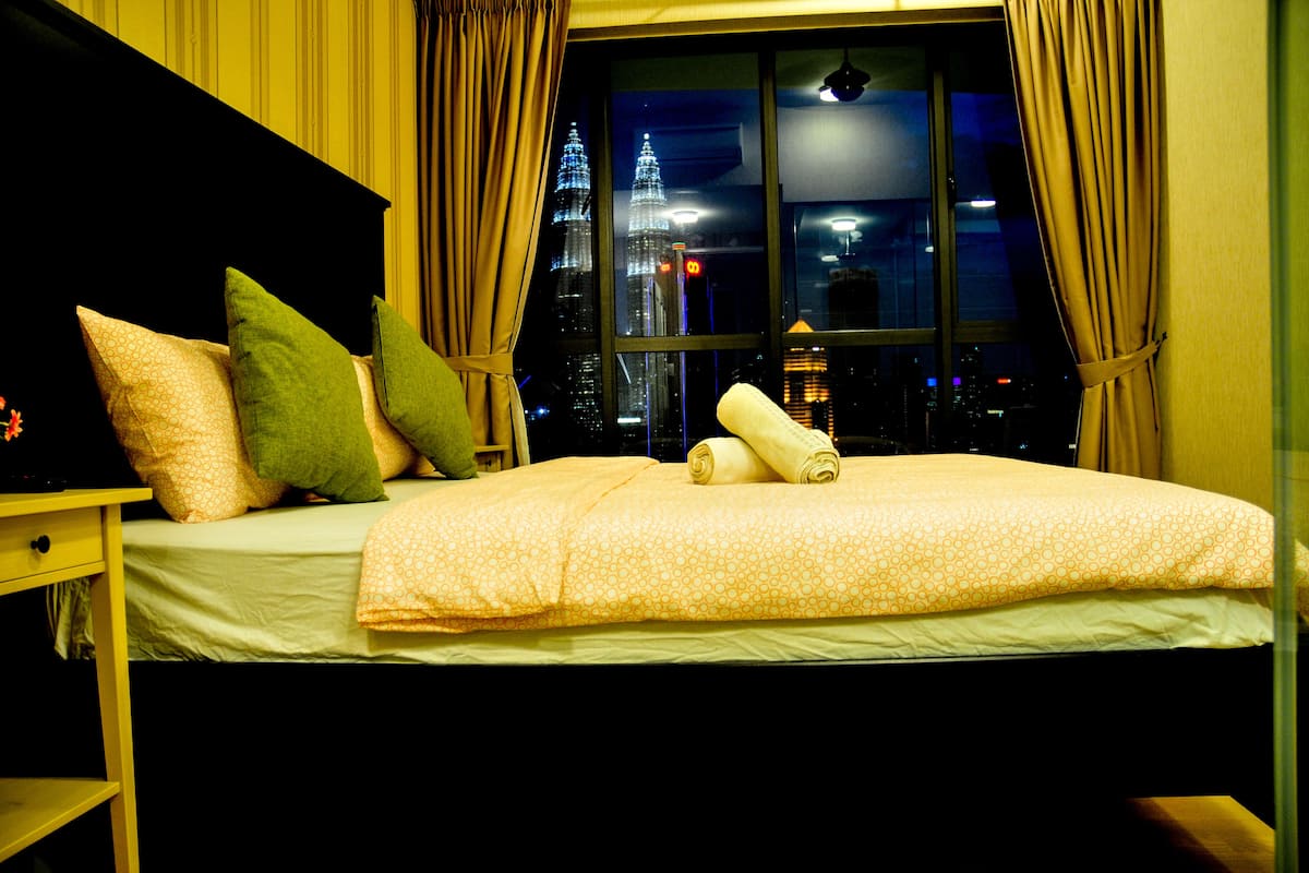 A neatly made bed with a patterned bedspread and pillows, a bedside table with a lamp, and a view of a cityscape through a window with curtains.