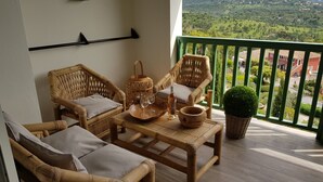 Varanda com móveis de vime, uma mesa com copos e uma garrafa, e vista para uma paisagem verde.