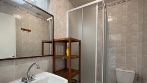 A bathroom with a white sink, a wooden shelf, a toilet, and a glass shower enclosure. - Studio Amiantis by Agence Bru - 2041 (Lacanau)