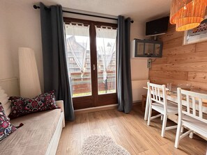 Una habitación con piso de madera, un sofá blanco con cojines estampados, una mesa de comedor con sillas y un ventanal con cortinas.