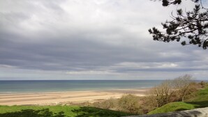 A coastal landscape with a stone wall in the foreground, a sandy beach, and a view of the ocean under a cloudy sky.
