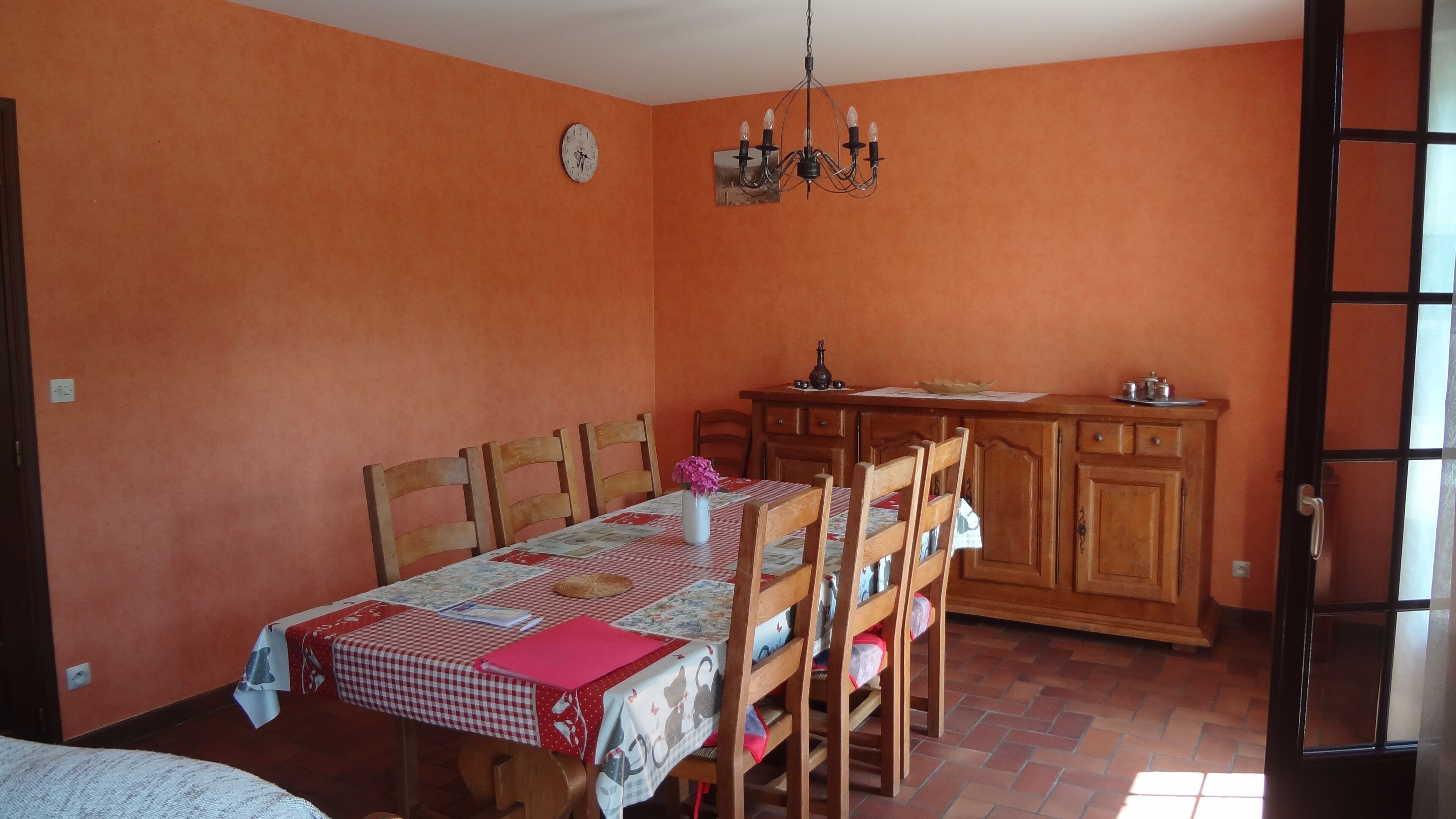 A dining area with a table set for four, wooden chairs, a chandelier, and a kitchen area with cabinets and a sink.