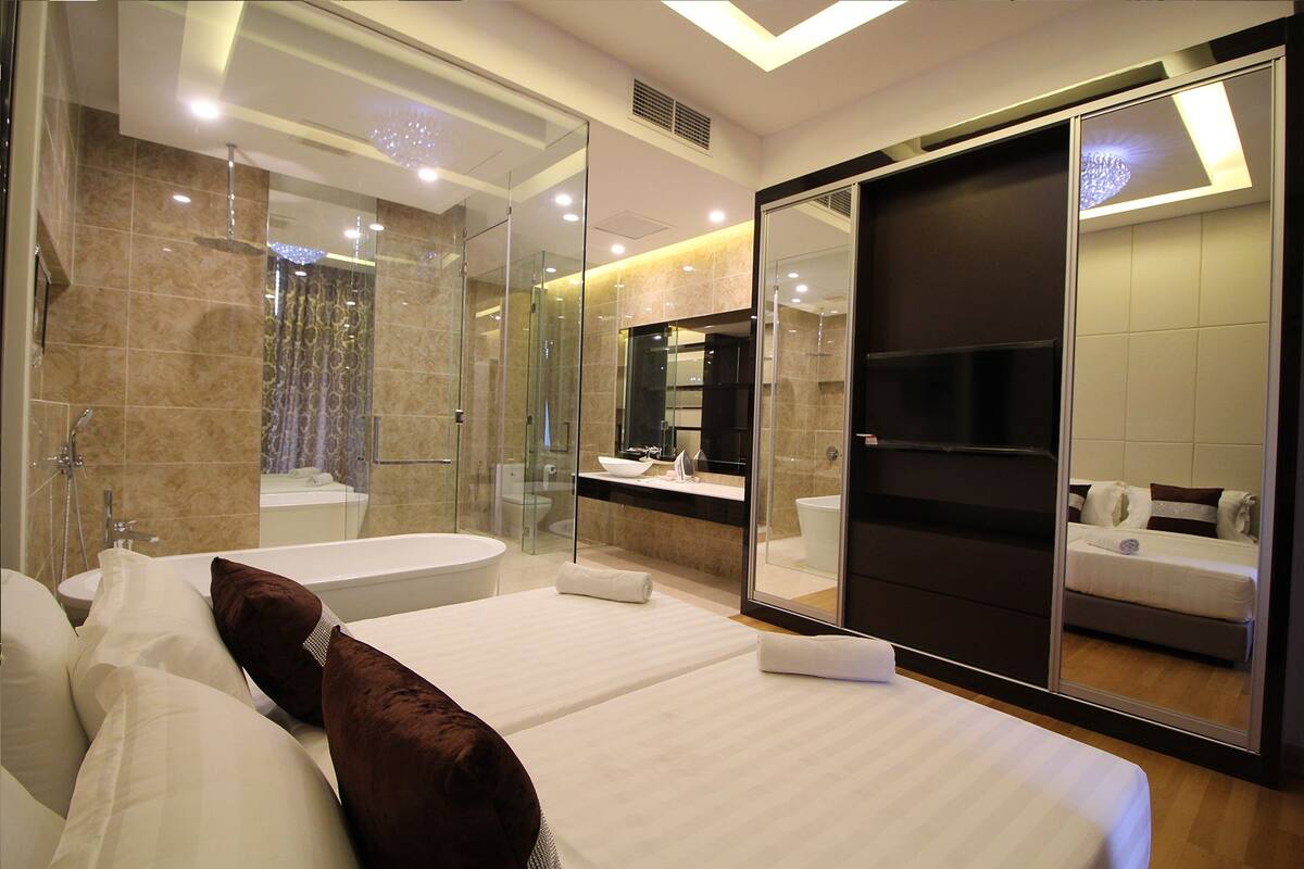 A modern hotel room with a large bed, a glass-enclosed shower, a large mirror, and a separate bathroom with a sink and toilet.