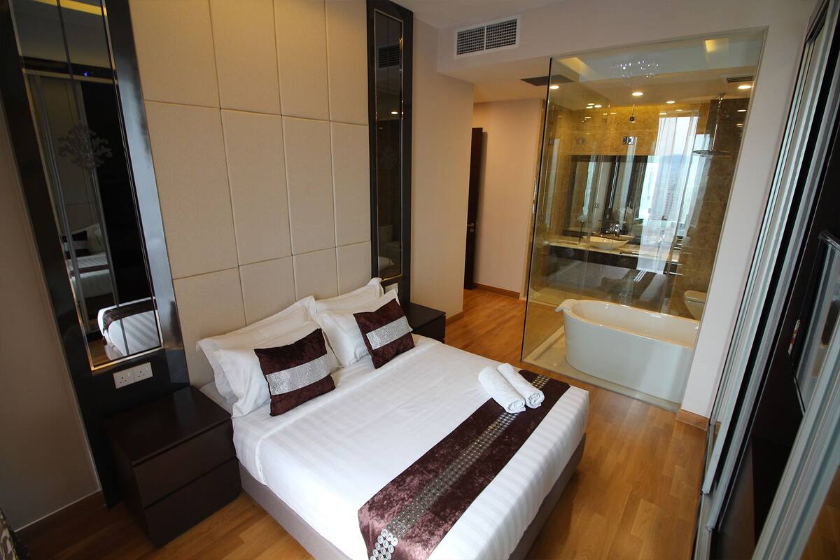A hotel room with a large bed, a bedside table, a mirror, and a glass-enclosed bathroom with a bathtub.