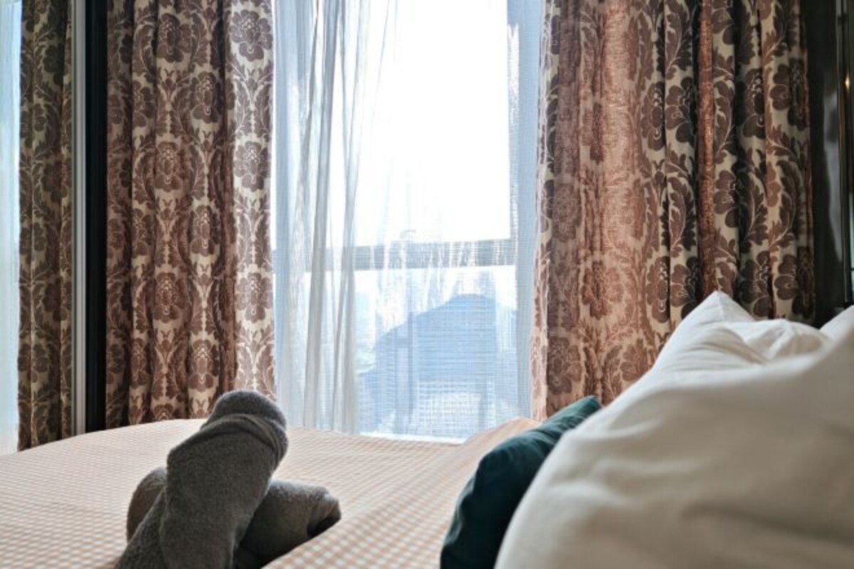 A bed with a checkered bedspread and a pillow, curtains with a floral pattern, and a view of a building through the window.
