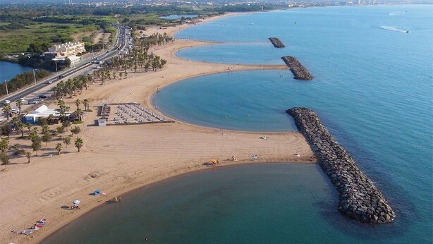 Vista aérea de una zona costera con playa de arena, una carretera con coches aparcados y un rompeolas rocoso que se adentra en el mar.