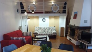 Una sala de estar con un sofá rojo, un sofá estampado, una mesa de comedor con una planta, un ventilador de techo y una chimenea.