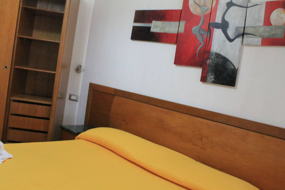 Quarto com cama, cabeceira de madeira e parede com arte abstrata.
