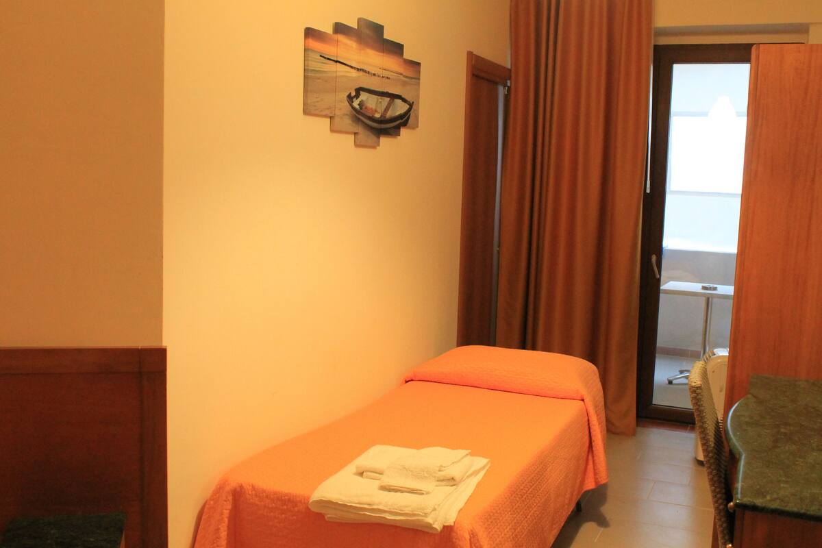 Quarto de hotel com cama, uma cadeira e porta que dá para uma varanda.