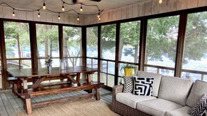 Une terrasse en bois avec une table à manger et des bancs, un canapé en osier agrémenté de coussins à motifs et de grandes fenêtres donnant sur une étendue d’eau.