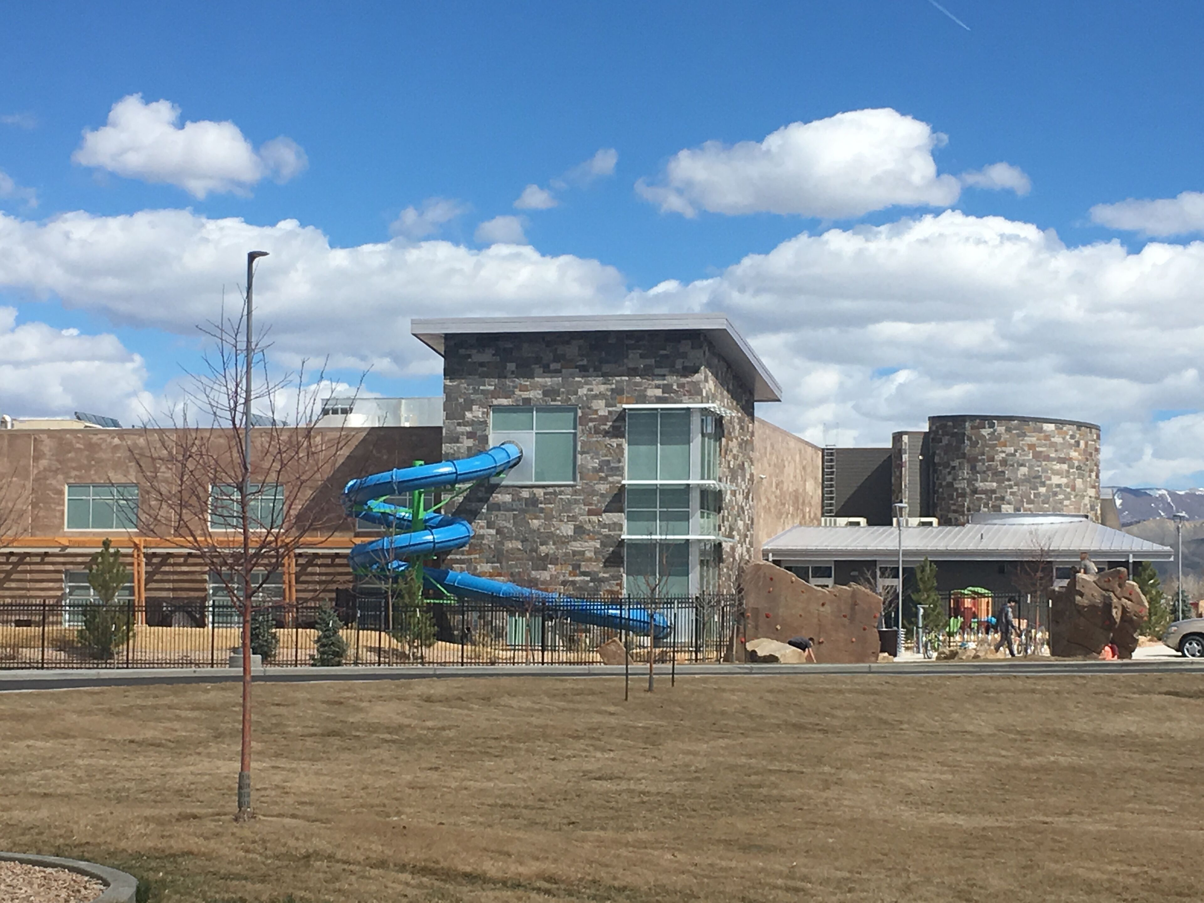 A playground with a blue slide and climbing wall, a stone building with large windows, and a clear blue sky.