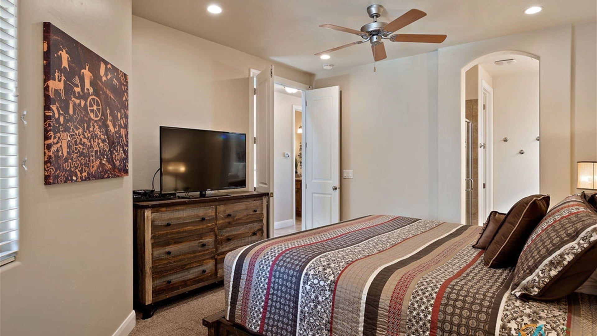 A bedroom with a bed, a television on a wooden dresser, a ceiling fan, and a door leading to another room.