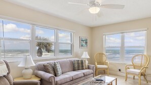 A living room with a beige sofa, two armchairs, a glass coffee table, and a ceiling fan. Large windows offer a view of the beach.