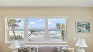 A room with a view of the beach through a large window, a couch with patterned cushions, and two bedside lamps.