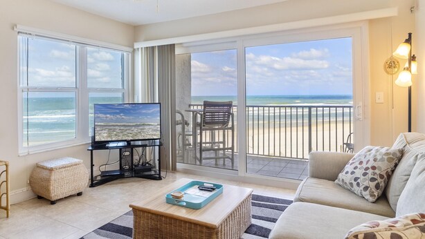 A living room with a large TV, a balcony with a chair, and a view of the beach.