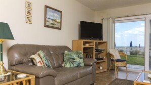 Ein Wohnzimmer mit einer Ledersofa, einem Fernseher, einem Bücherregal und einem Korbsessel.