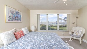 Ein Schlafzimmer mit einem großen Bett, einem Fenster mit Blick auf Grünflächen und Gebäude, einem Sessel mit einem herzförmigen Kissen und einem Deckenventilator.