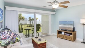A living room with a large TV, a ceiling fan, and a view of the ocean through the sliding glass doors.