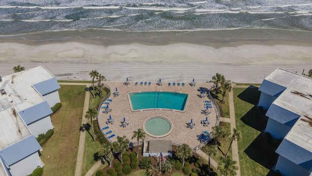 An aerial view of a beachfront hotel with a swimming pool, beach chairs, and palm trees.