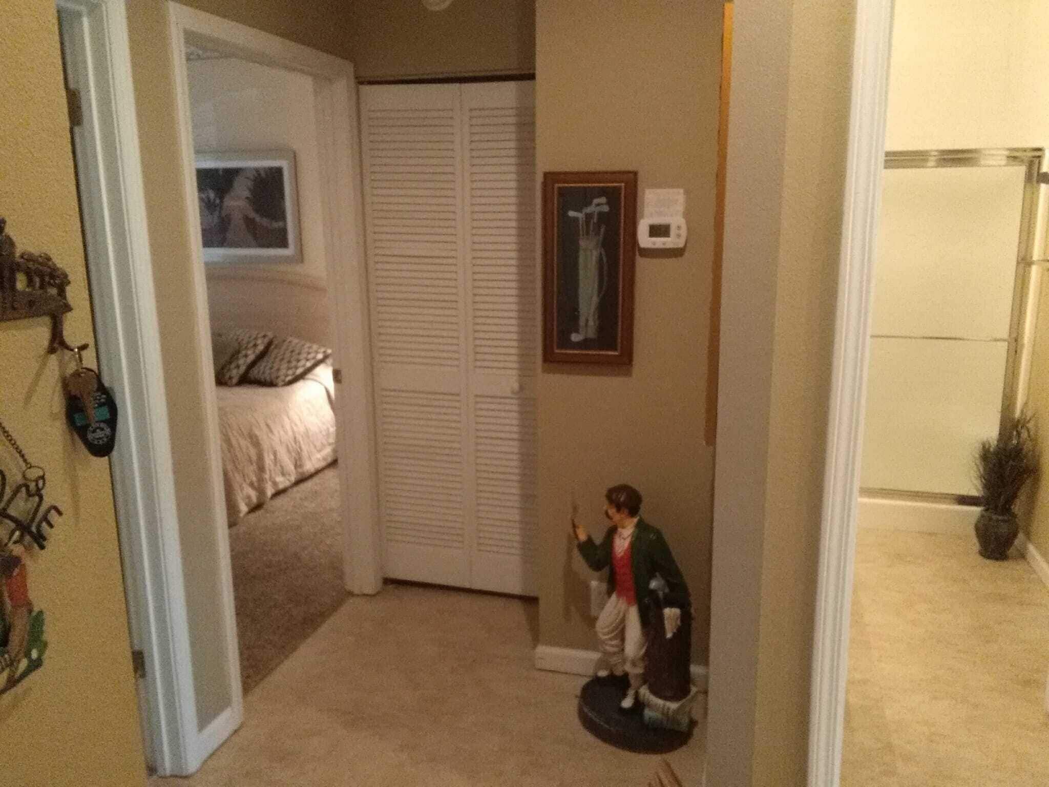 A hallway with a bed in the background, a coat rack with items, and a framed picture on the wall.