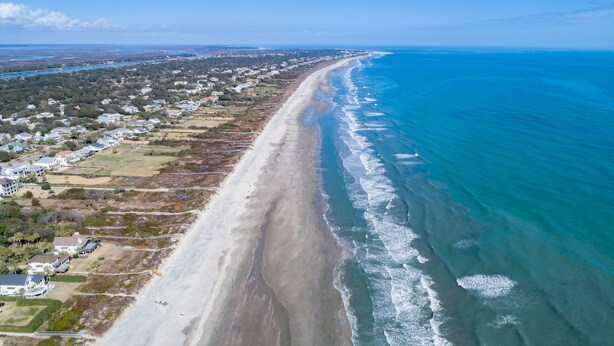 Vista aérea de una zona costera con una extensa playa de arena, propiedades residenciales y un océano azul cristalino.