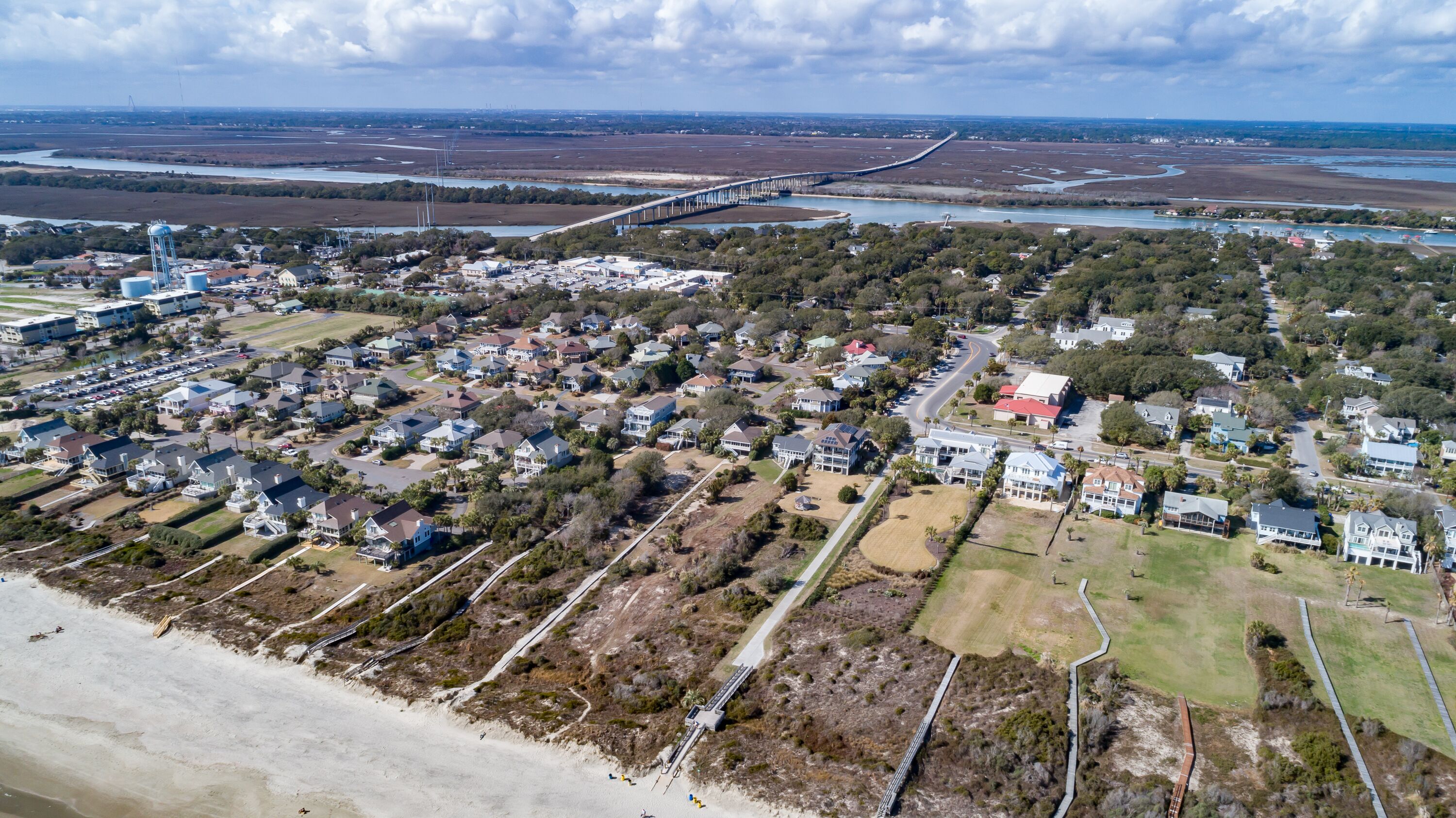 An aerial view of a coastal residential area with houses, a beach, and a bridge in the background.